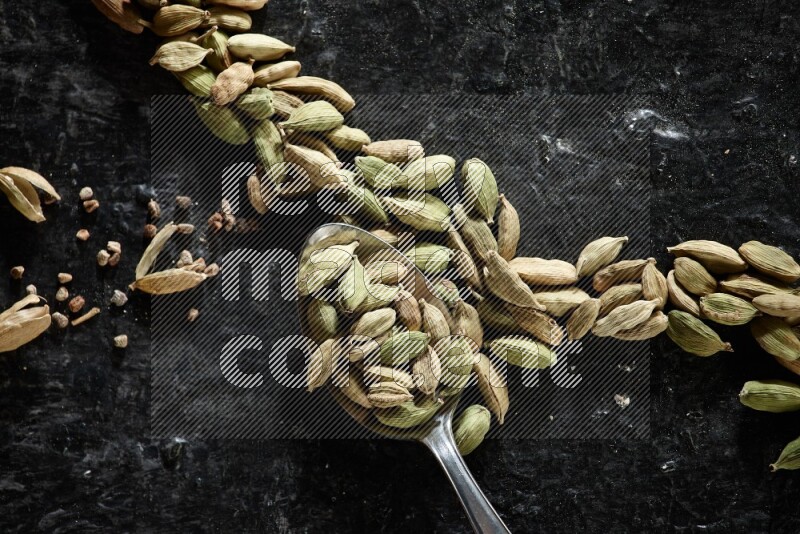 A Metal spoon full of cardamom seeds and some seeds beside it on a textured black flooring