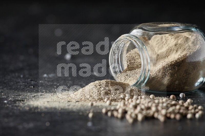 A flipped herbal glass jar full of white pepper powder with spilled powder and pepper beads on textured black flooring