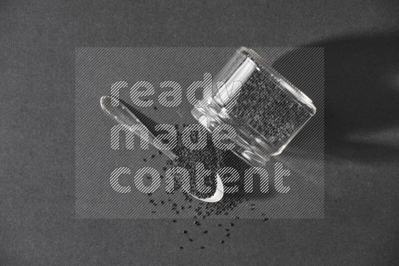 A flipped glass jar full of black seeds and seeds spread out with a metal spoon full of the seeds on a black flooring