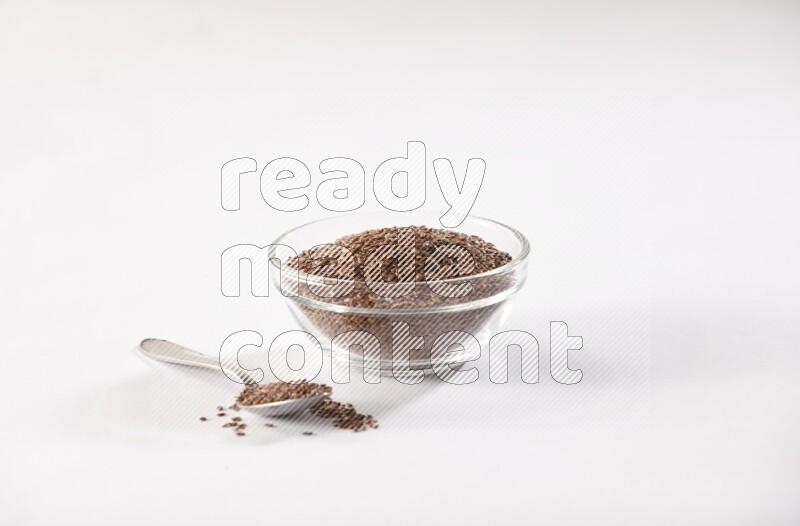 A glass bowl full of flax seeds and a metal spoon full of the seed on a white flooring