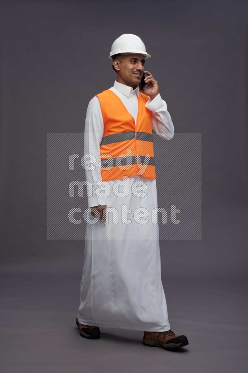 Saudi man wearing thob with engineer vest standing talking on phone on gray background