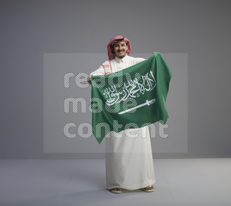 A saudi man standing wearing thob and red shomag holding big saudi flag on gray background