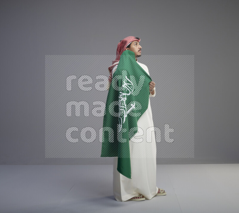 A saudi man standing wearing thob and red shomag holding big saudi flag on gray background