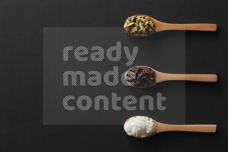 الفلفل الأبيض والقرنفل والهيل والملح والفلفل الأسود وريحان مجفف في معالق خشبية علي خلفية سوداء