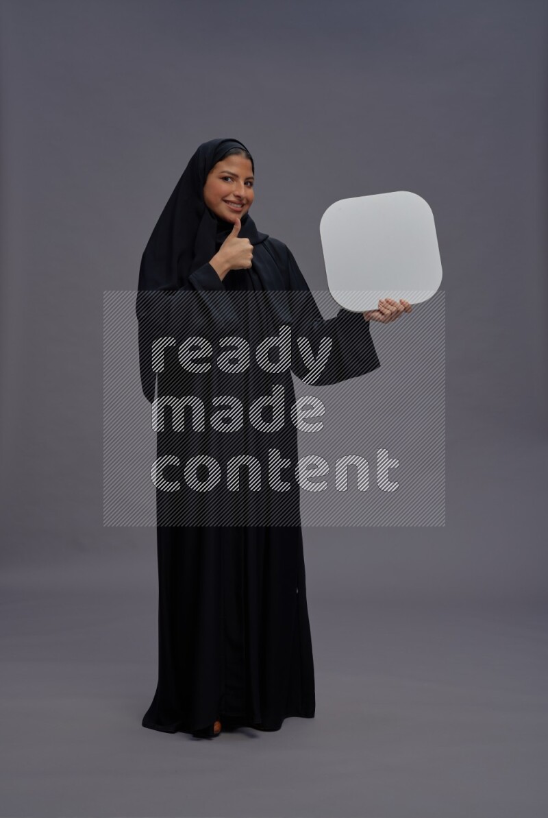 Saudi woman wearing Abaya standing holding social media sign on gray background