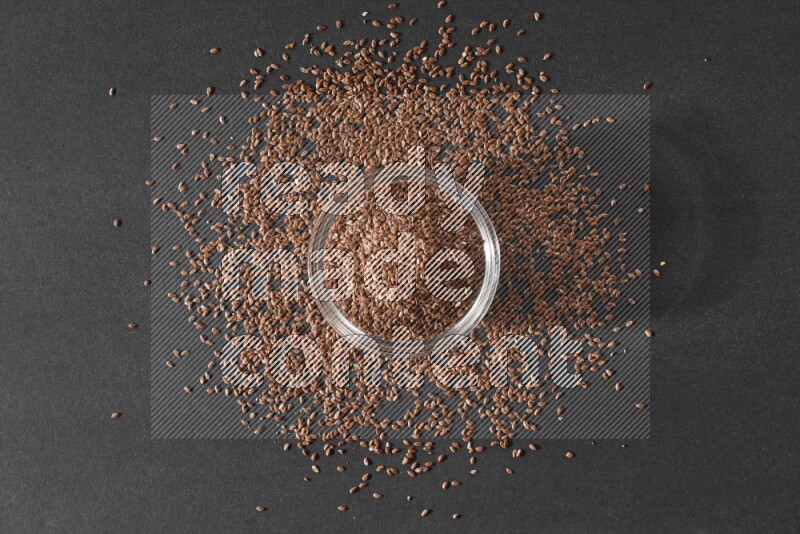 A glass bowl full of flaxseeds surrounded by the seeds on a black flooring