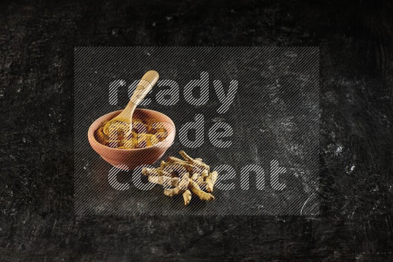 A wooden bowl and a wooden spoon full of turmeric powder with dried turmeric fingers on textured black flooring