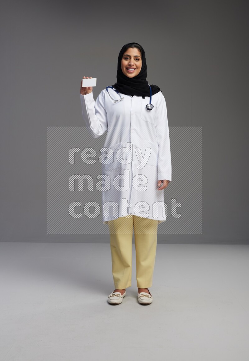 Saudi woman wearing lab coat with stethoscope standing holding ATM card on Gray background
