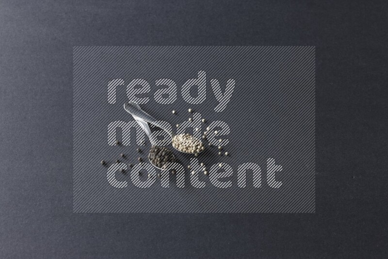 ملعقتان معدنيتان ممتلئتان بحبوب الفلفل الأسود والأبيض مع حبوب متناثرة على أرضية سوداء