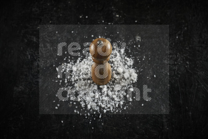 A wooden grinder standing upright and surrounded by coarse white sea salt on black background