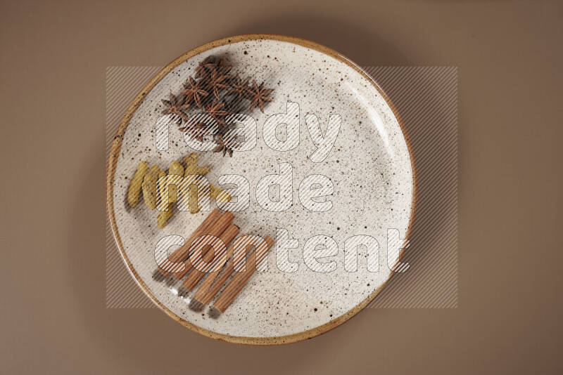 An assorted whole spices on a flat beige plate including fresh and dried ginger, cinnamon sticks, cardamom, turmeric fingers, cloves, star anise on a beige background