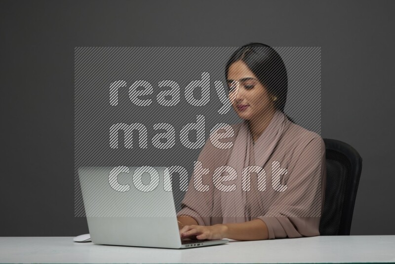 A Saudi woman Sitting on her desk on a Gray Background wearing Brown Abaya