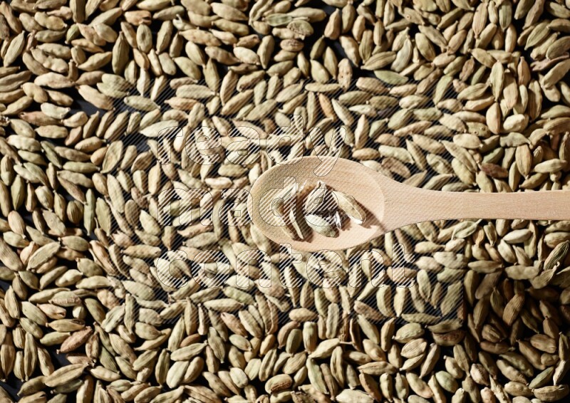Cardamom seeds in a wooden spoon above cardamom seeds background on black flooring