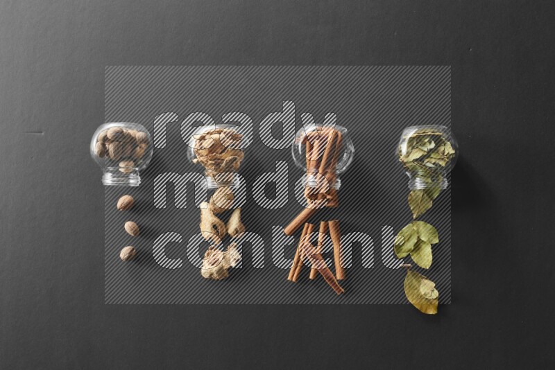 4 glass spice jars full of laurel bay leaves, ginger, cinnamon and nutmeg on black flooring
