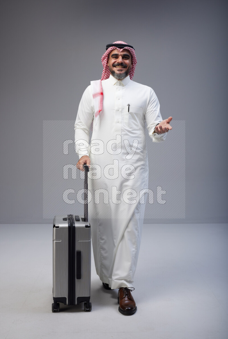Saudi man wearing Thob and red Shomag standing holding Travel bag on Gray background