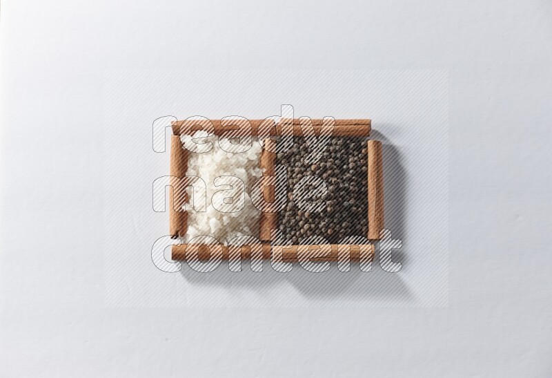 مربعان من أعواد القرفة ممتلئان بحبوب الفلفل الأسود والملح الأبيض علي خلفية بيضاء