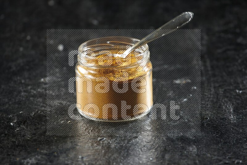 A glass jar and a metal spoon full of turmeric powder on a textured black flooring