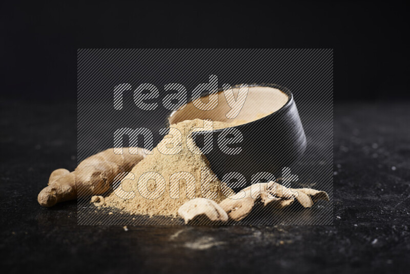 A black pottery bowl full of ground ginger powder with fallen powder from it on black background