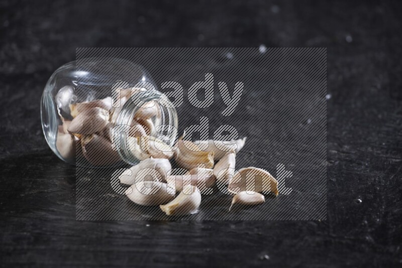 A glass spice jar full of garlic cloves flipped over with the cloves spilling out on a textured black flooring
