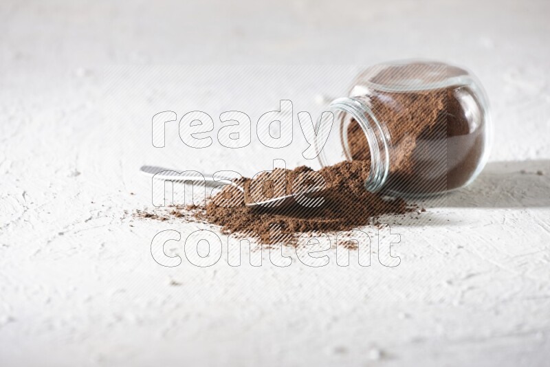 A flipped glass spice jar and a metal spoon full of cloves powder and powder came out of the jar on textured white flooring