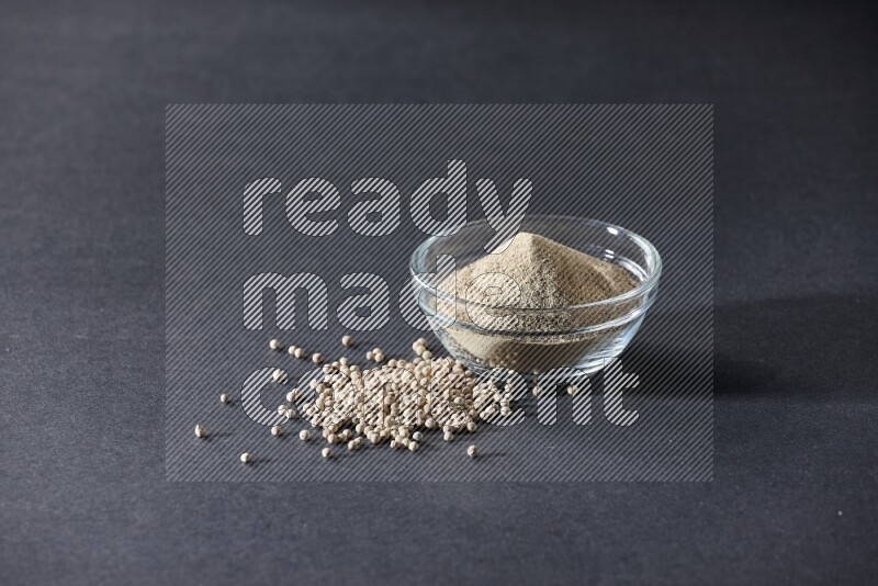 A glass bowl full of white pepper powder with white pepper beads on black flooring
