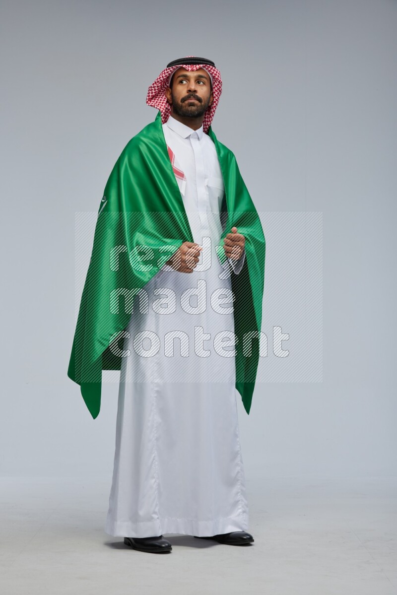 Saudi man Wearing Thob and shomag standing holding Saudi flag on Gray background