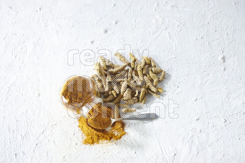 وعاء زجاجي للتوابل مقلوب وملعقة معدنية ممتلئة ببودرة الكركم والبودرة تناثرت منه مع أصابع الكركم المجففة على خلفية بيضاء