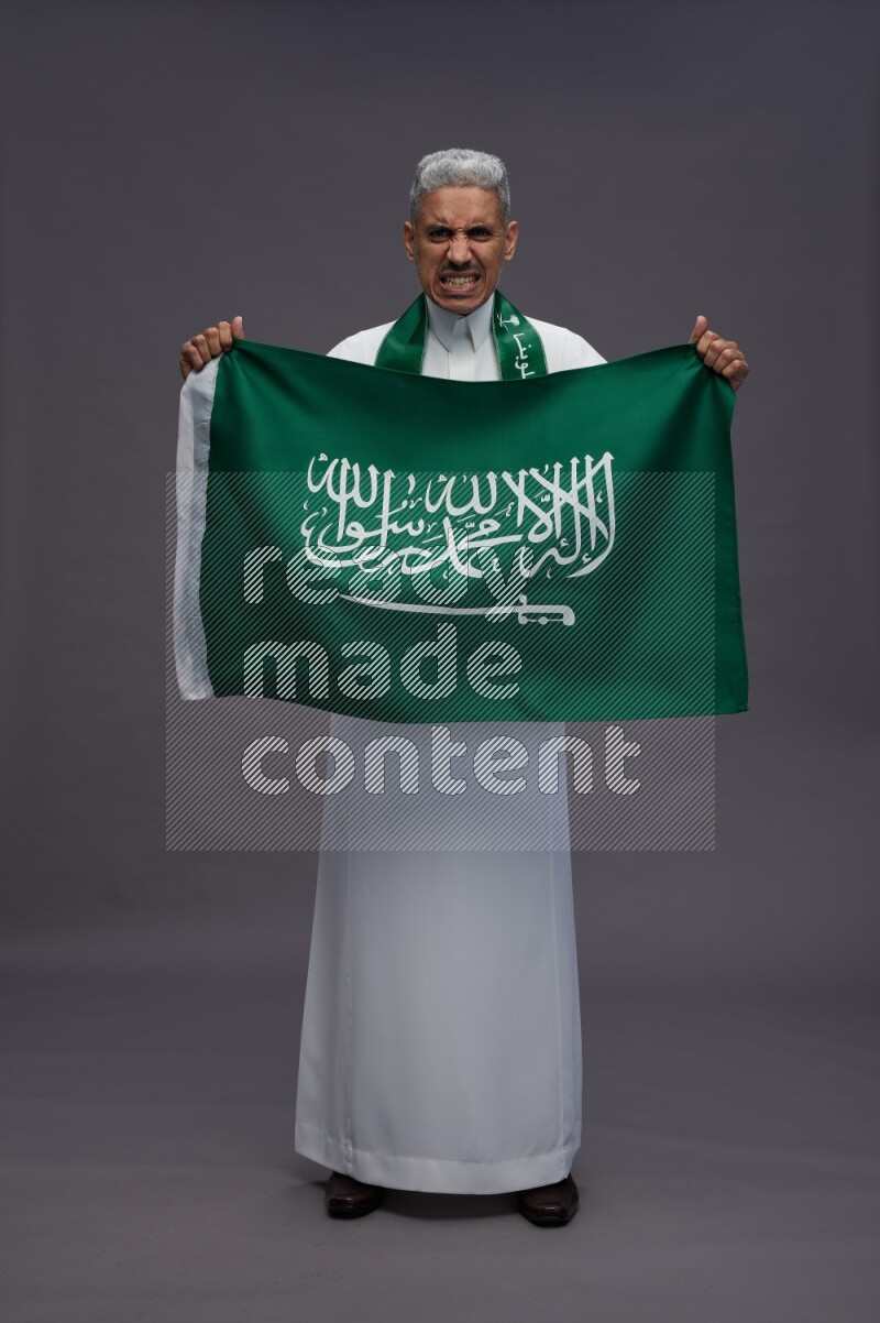 Saudi man wearing thob standing holding Saudi flag on gray background