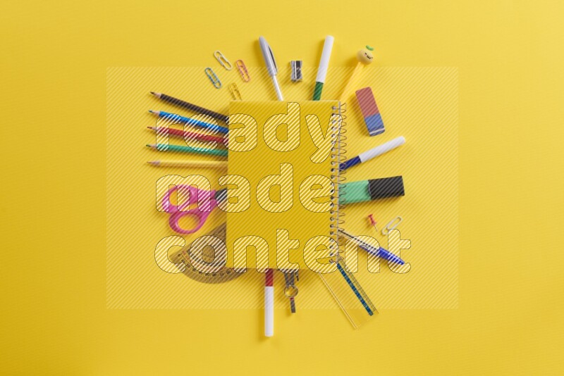 A yellow notebook with different school supplies on yellow background