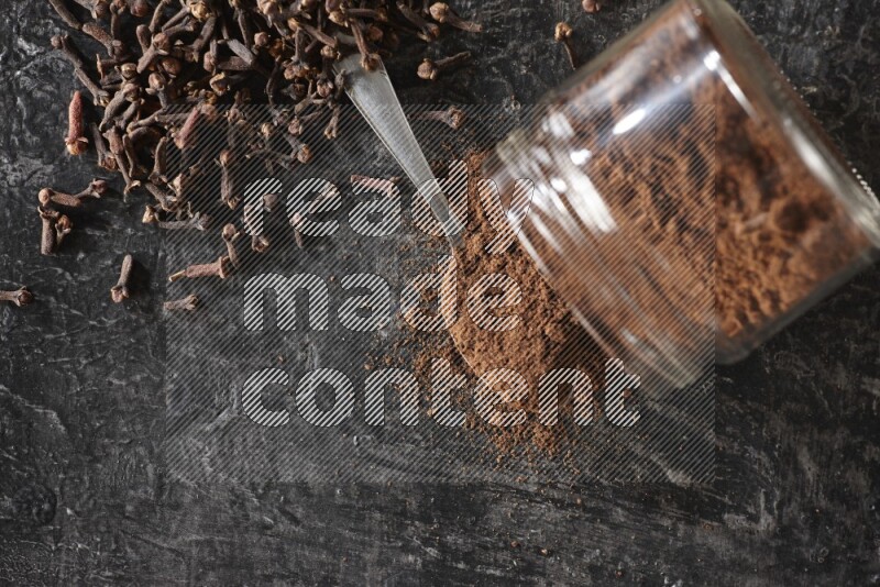 A flipped glass jar and metal spoon full of cloves powder with cloves spread on a textured black flooring