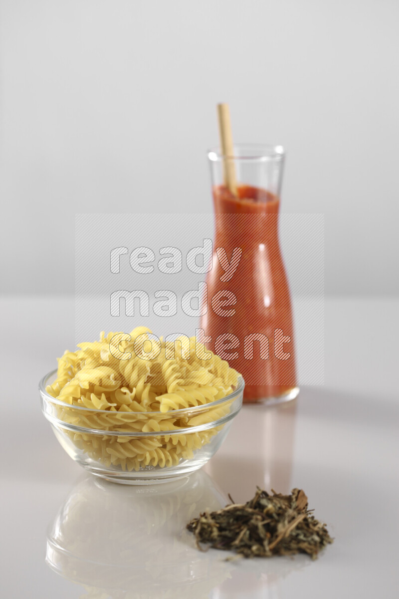 Raw pasta with tomatoe pasta with different ingredients such as cherry tomatoes, basil, garlic, bay laurel, cardamom, white pepper, black pepper, red chilis and wheat stalks on light grey background