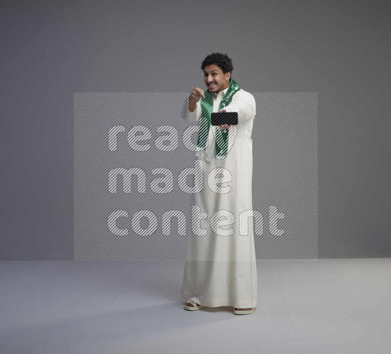 A Saudi man standing wearing thob and Saudi flag scarf showing phone to camera on gray background