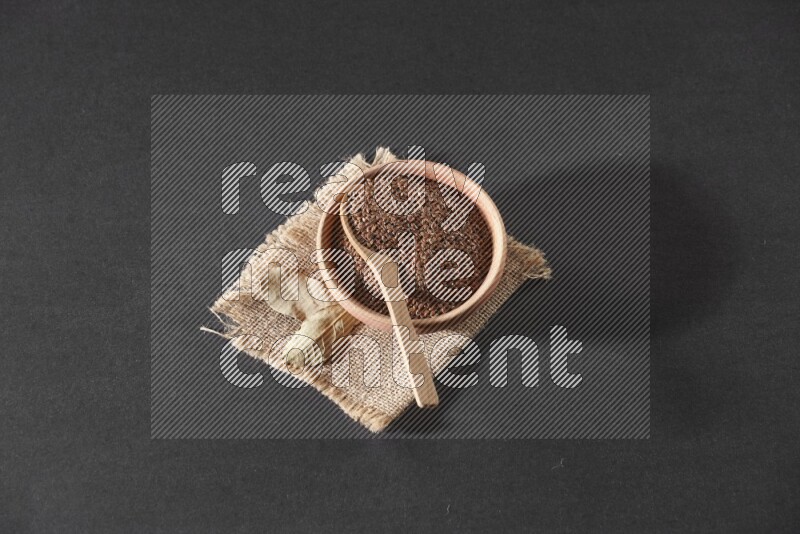 وعاء خشبي ممتلئ بحبوب بذر الكتان مع ملعقة خشبية ممتلئة بالحبوب علي قطعة من القماش علي خلفية سوداء