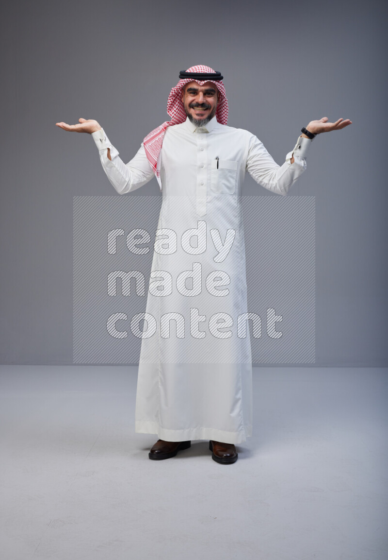 Saudi man Wearing Thob and red white Shomag standing interacting with the camera on Gray background