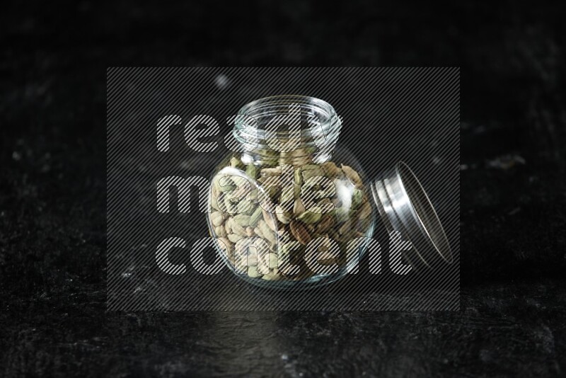 A glass spice jar full of cardamom seeds on textured black flooring