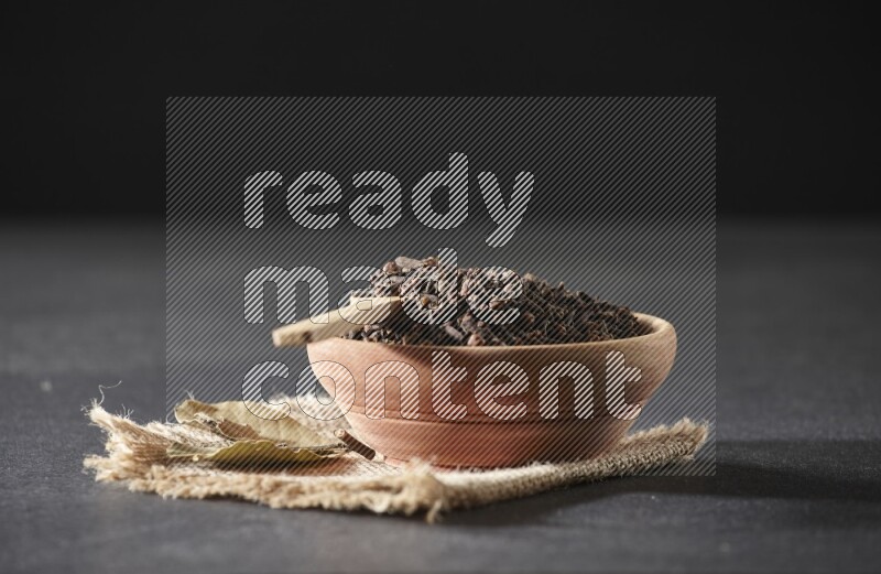 A wooden bowl, a wooden spoon full of cloves, and bay leaves (laurel) on a piece of burlap on a black flooring