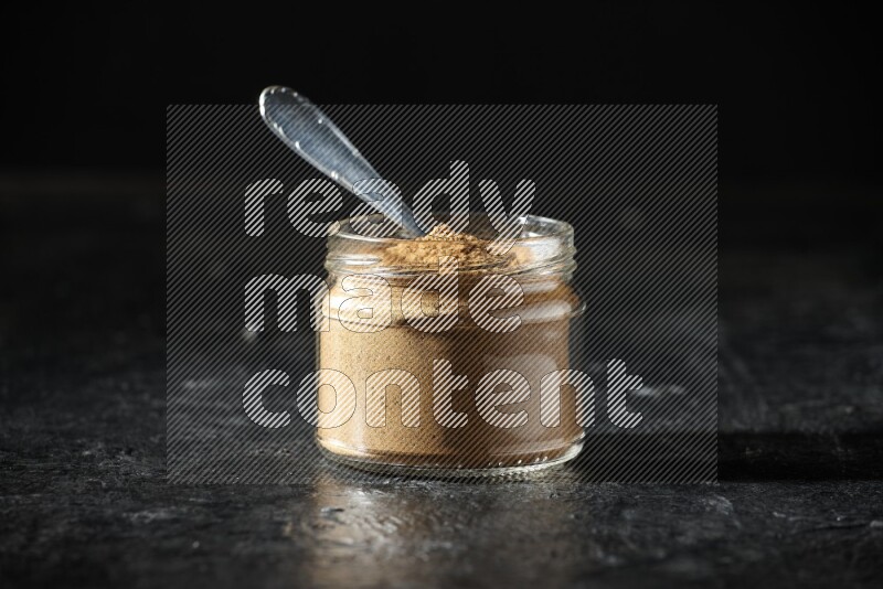 A glass jar and a metal spoon full of allspice powder on a textured black flooring