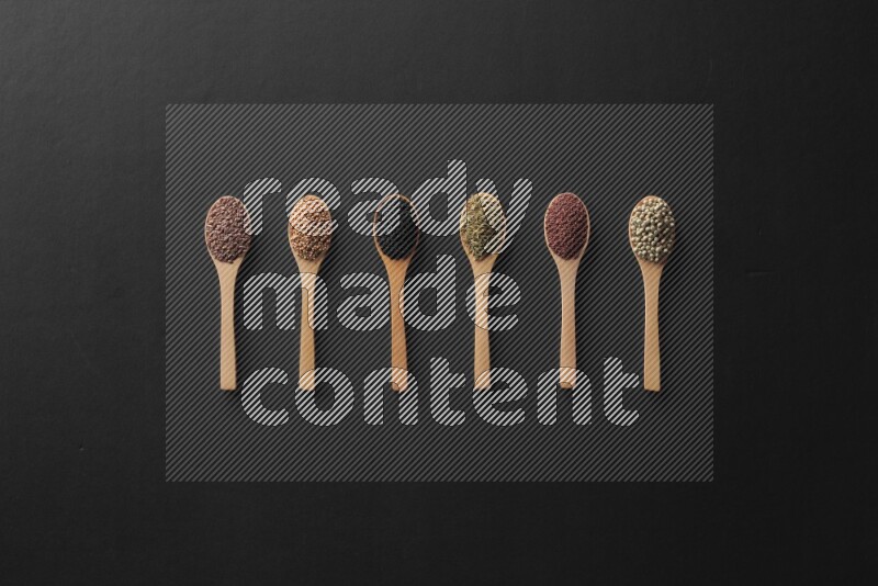 wooden spoons filled with white peppers, mustard seeds, black seeds, garden cress, cumin and flax on black flooring