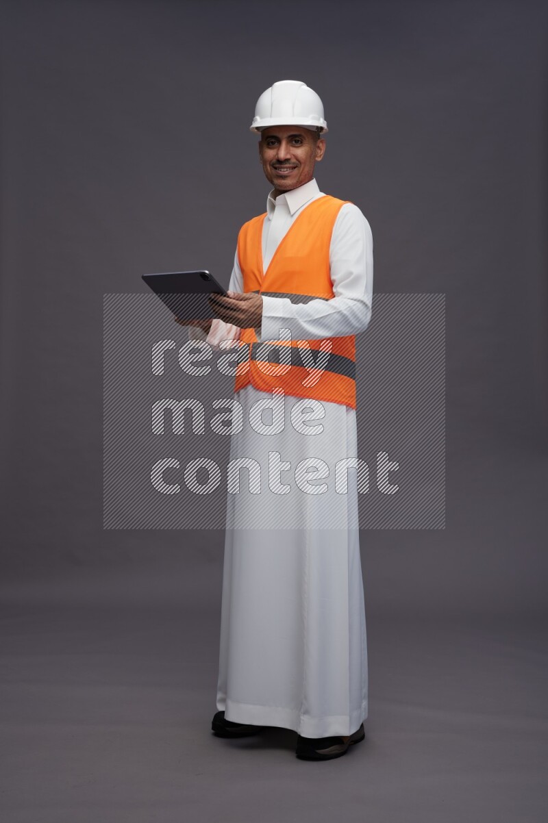 Saudi man wearing thob with engineer vest standing working on tablet on gray background