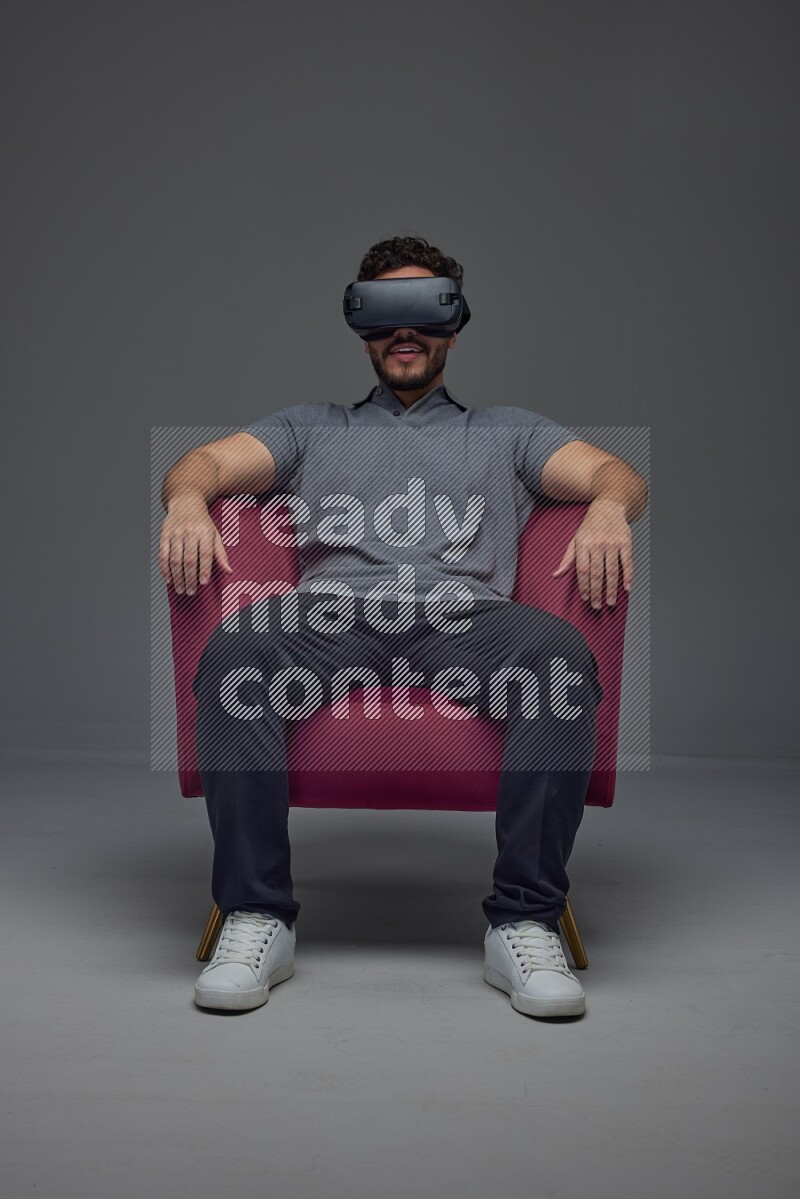 A man wearing casual and wearing VR while sitting on a burgundy chair eye level on a gray background