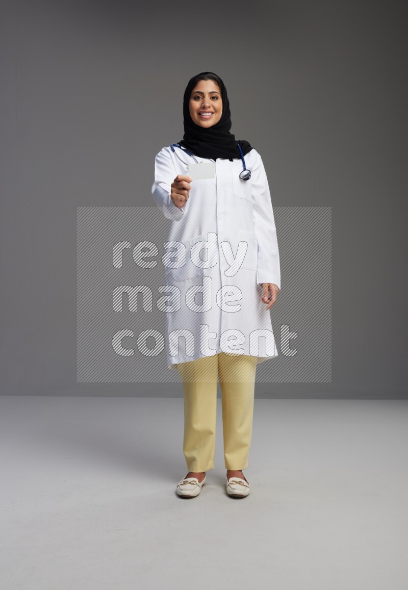Saudi woman wearing lab coat with stethoscope standing holding ATM card on Gray background
