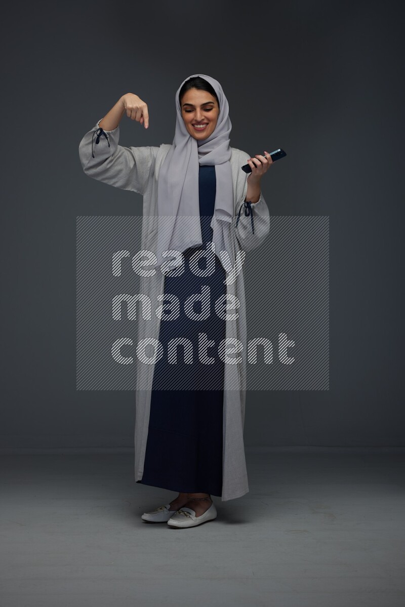 A Saudi woman wearing a light gray Abaya and head scarf standing and holding a phone on a grey background