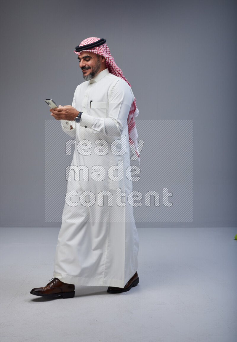 Saudi man Wearing Thob and red Shomag standing texting on phone on Gray background