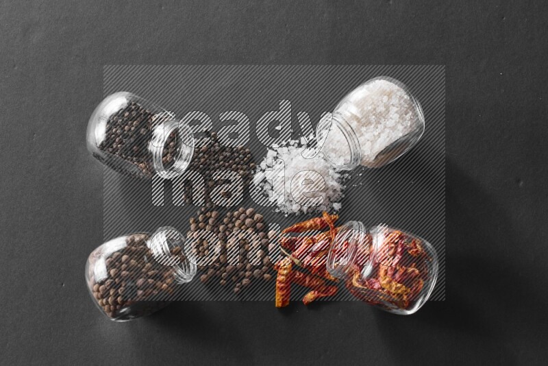 4 glass spice jars full of salt, chili peppers, black peppers and allspice on black flooring