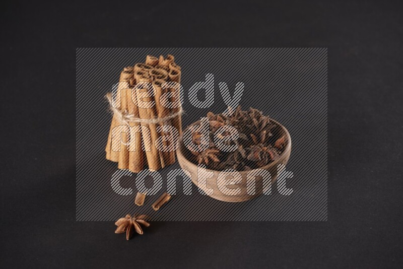 A stacked and bounded cinnamon sticks and a wooden bowl full of star anise on a black background
