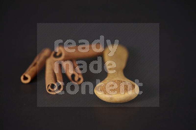 Cinnamon powder in a wooden spoon with cinnamon sticks beside it on black background