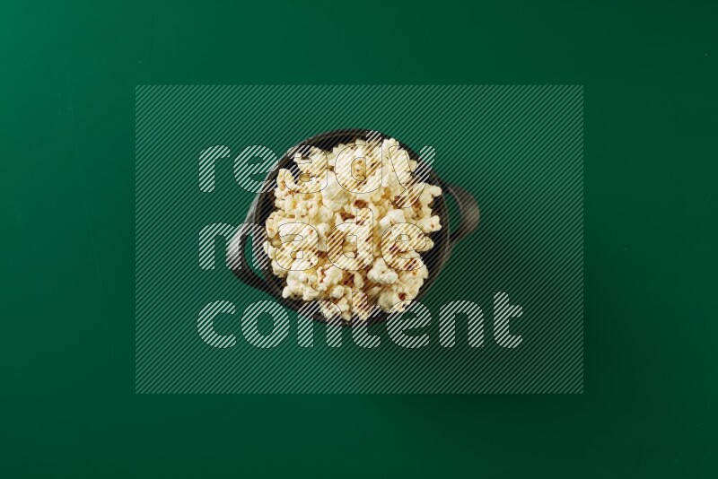 A black ceramic bowl full of popcorn on a green background in a top view shot
