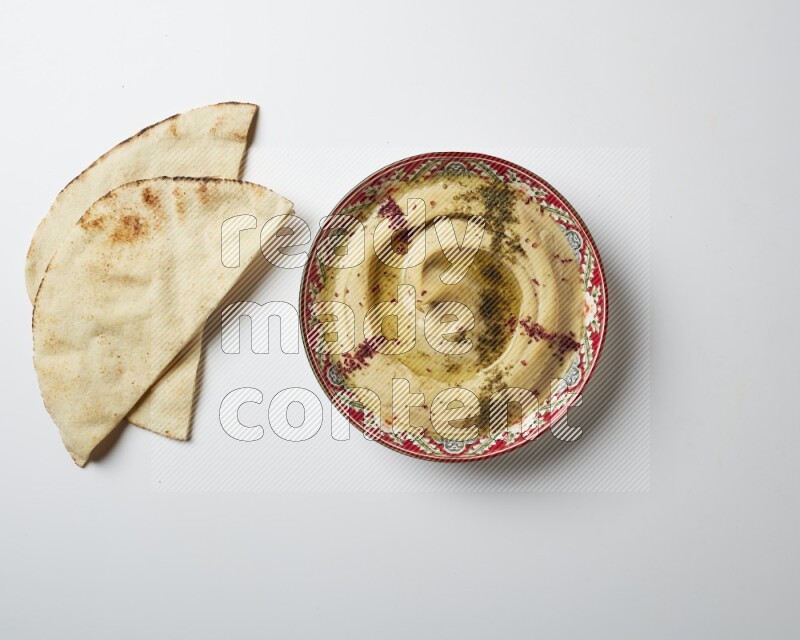Hummus in a red plate with patterns garnished with zaatar & sumak on a white background