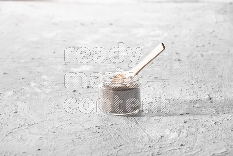 A glass jar full of black pepper powder with a wooden spoon on a textured white flooring
