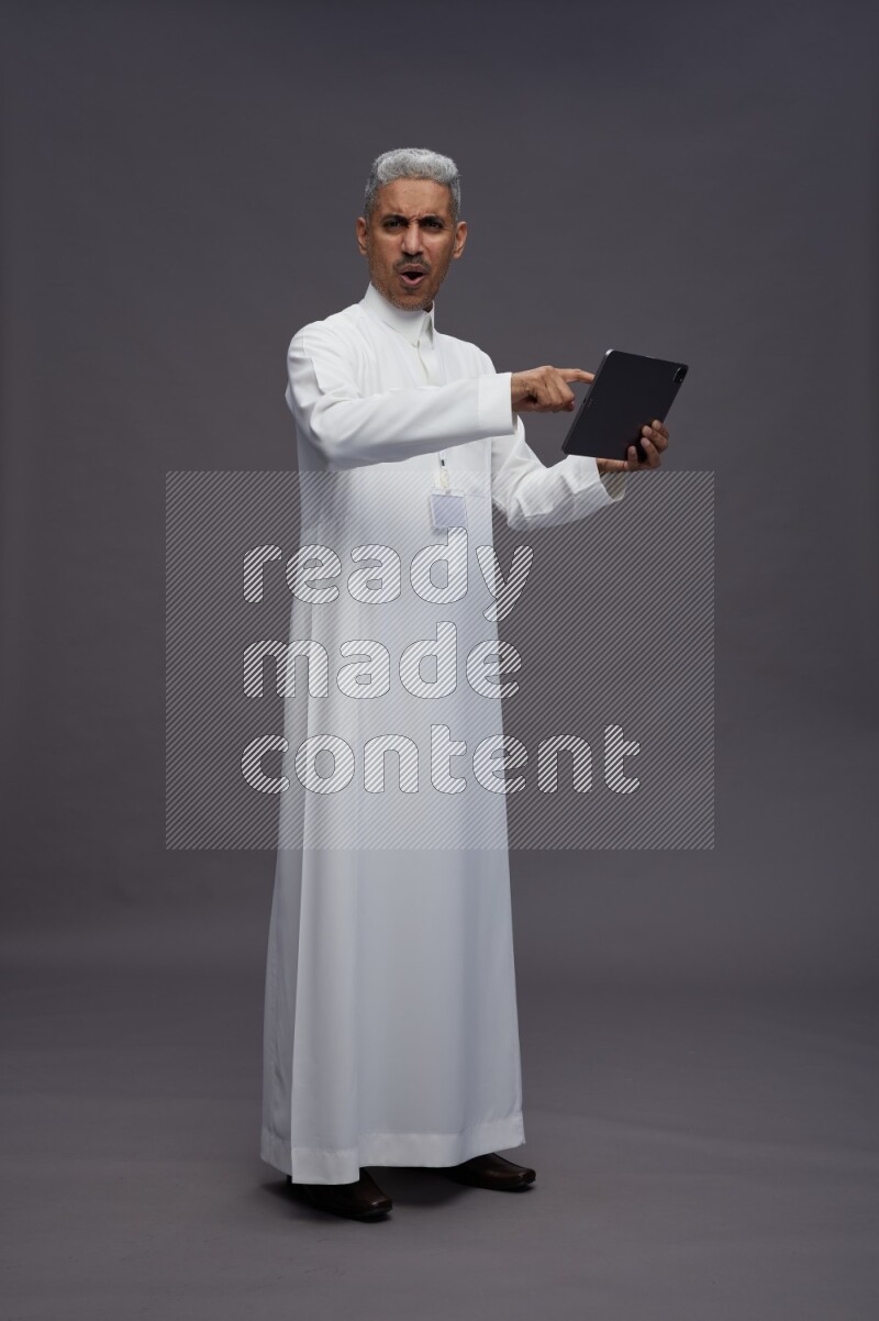 Saudi man wearing thob with neck strap employee badge standing working on tablet on gray background